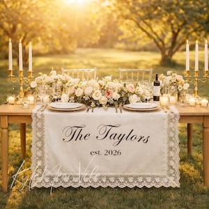 Puede incluir: Una mesa de boda bellamente decorada con un camino de mesa de encaje color crema. El camino de mesa lleva impreso "The Taylors est. 2026". La mesa está adornada con flores, velas y detalles dorados. La escena está ambientada al aire libre.