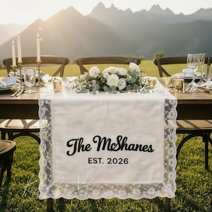 May include: A cream-colored table runner with a delicate lace trim, featuring the text "The McShanes EST. 2026" in elegant script. The runner is set on a wooden table, adorned with floral arrangements and candles, creating a romantic ambiance.