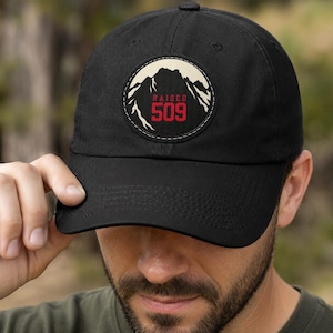 May include: A black baseball cap featuring a circular patch on the front. The patch displays a cream-colored mountain silhouette against a black background, with the red text RAISED 509 centered below the mountain peak. The cap has a curved brim.