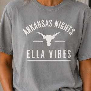 Puede incluir: Una camiseta gris de manga corta con texto impreso en blanco que dice ARKANSAS NIGHTS y ELLA VIBES, separado por un gráfico blanco de un cráneo de toro longhorn. La camiseta tiene cuello redondo y un corte estándar.