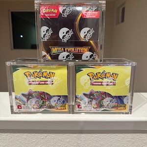 Puede incluir: Tres vitrinas transparentes de acrílico que contienen cajas de cartas coleccionables de Pokémon. La vitrina superior muestra una caja con el texto "MEGA EVOLUTION". Las vitrinas inferiores contienen cajas con el texto "EVOLVING SKIES" y la marca Pokémon.