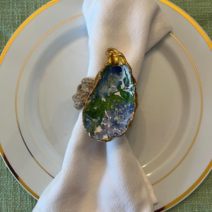 May include: A white linen napkin is held by a decorative oyster shell napkin ring. The shell is painted with blue and green floral designs and trimmed with gold. The napkin and ring rest on a white plate with a gold rim.