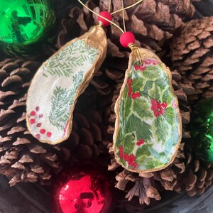 May include: Two gold-painted oyster shell Christmas ornaments. One ornament features a green and white holly pattern with red berries. The other ornament features a green and white pine tree pattern with red berries.