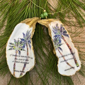 May include: Two oyster shell ornaments with gold edges, featuring palm tree designs and quotes by Jimmy Buffett. One says, "If there's a heaven for me, I'm sure it has a beach attached." The other says, "No working during drinking hours."