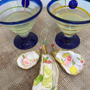 May include: Three gold-painted oyster shell ornaments with painted images of cocktails. The ornaments feature a pink cocktail with a lime wedge, a green cocktail with a lime wedge, and a pink cocktail with a cherry and a lime wedge.