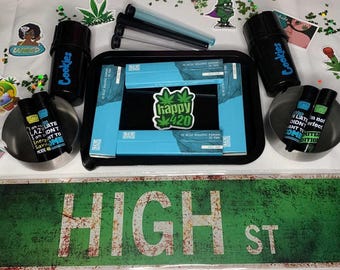 Può includere: Un cartello stradale verde e bianco con la scritta "HIGH ST" su di esso. Il cartello è circondato da vari oggetti, tra cui un vassoio nero con un imballaggio blu e bianco, un contenitore nero con la scritta "Cookies" su di esso e due piccoli contenitori blu e bianchi con le parole "Sono così pigro che non voglio nemmeno fumare erba" su di essi.