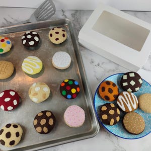 May include: A collection of colorful, felt cookies arranged on a metal baking sheet and a blue plate. The cookies feature various designs, including polka dots, stripes, and textured patterns. A white bakery box with a clear window is also present.