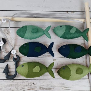 May include: A decorative mobile featuring six felt fish in shades of green and blue, suspended from a wooden rod. The mobile includes two felt hooks and a gray ribbon for hanging. The fish are approximately 4 inches long.