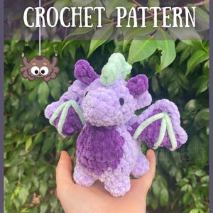 以下が含まれることがあります： 紫と緑の色合いの手作りの編み物ドラゴン。テキスト「OPAL THE DRAGON CROCHET PATTERN」と「@cobwebcrochet」があります。ドラゴンは翼とふわふわした質感があり、緑の葉の背景に対して手で持たれています。