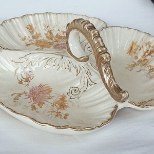 Puede incluir: Un plato de servir de cerámica blanca con tres secciones y un asa dorada. El plato está decorado con un patrón floral en tonos marrones, rosas y dorados.