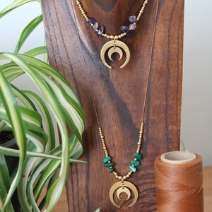May include: Two gold necklaces with crescent moon pendants. The top necklace features amethyst beads and the bottom necklace features malachite beads.