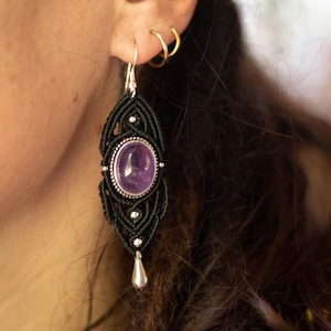 May include: A black macramé earring featuring an oval amethyst gemstone and a silver teardrop charm. The earring hangs from a silver hook. The earring is worn on a person's ear.