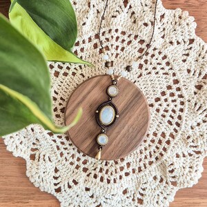 May include: A macrame necklace featuring three oval moonstone cabochons set in dark brown cord. The central stone is the largest, flanked by two smaller stones, with a small gold-toned metal teardrop charm at the bottom and small beads on the cord.