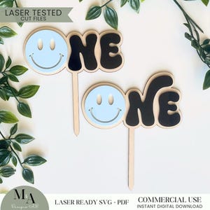 Pode incluir: Dois toppers de bolo de madeira cortados a laser com a palavra "ONE" em preto e um rosto sorridente azul. O texto "LASER READY SVG + PDF" e "COMMERCIAL USE INSTANT DIGITAL DOWNLOAD" está na parte inferior da imagem.