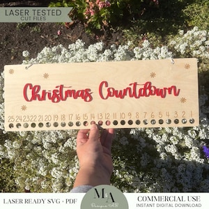 Puede incluir: Un letrero de cuenta atrás de Navidad de madera con 25 círculos para contar los días hasta Navidad. El letrero está pintado con texto rojo que dice "Christmas Countdown" y tiene copos de nieve dorados en la parte superior e inferior.