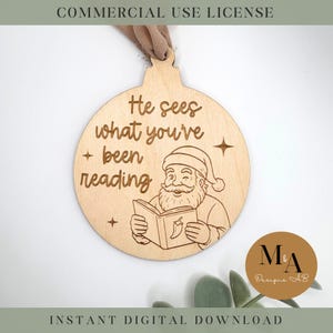 May include: Wooden Christmas ornament with the text "He sees what you've been reading" and an illustration of Santa Claus reading a book. The ornament has a light brown color and a loop for hanging.