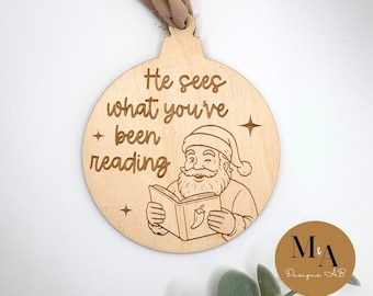 May include: Wooden Christmas ornament with the text "He sees what you've been reading" and an illustration of Santa Claus reading a book. The ornament has a light brown color and a loop for hanging.