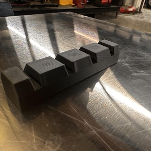 May include: A black plastic tool with four rectangular blocks, each with a textured surface. The tool is designed for holding and organizing small objects.