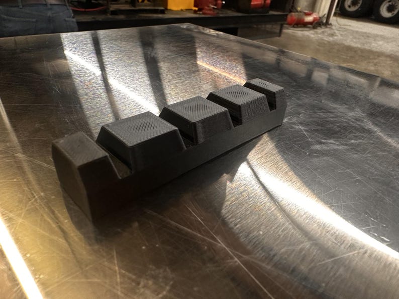 May include: A black plastic tool with four rectangular blocks, each with a textured surface. The tool is designed for holding and organizing small objects.