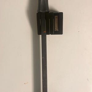 Puede incluir: Una palanca negra con un mango texturizado y un extremo plano y biselado. La herramienta está montada sobre una superficie blanca con dos soportes negros. Una pequeña placa de metal está adherida a uno de los soportes.