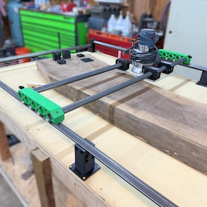 Puede incluir: Una máquina enrutadora CNC con una pieza de trabajo de madera. La máquina tiene un motor azul y negro, soportes verdes y un marco de metal. La pieza de trabajo es un trozo grande y rectangular de madera marrón.