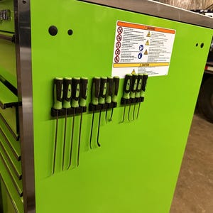 Puede incluir: Una caja de herramientas de metal verde brillante con varios cajones. La caja de herramientas tiene una tira magnética en el lateral con una variedad de herramientas, incluyendo destornilladores y barras de palanca. La caja de herramientas tiene una etiqueta de advertencia con instrucciones de seguridad.