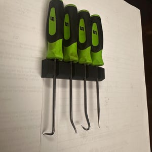 May include: Four black and green handled tools with metal hooks hang on a black wall mount. The tools are labeled "LS".