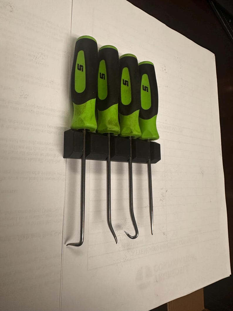 May include: Four black and green handled tools with metal hooks hang on a black wall mount. The tools are labeled "LS".