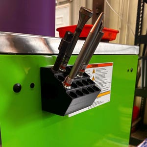 May include: A black, angled tool holder with multiple cylindrical openings, mounted on a bright green surface. Various tools, including metal rods and cylindrical components, are inserted into the holder. A warning label is visible on the green surface.