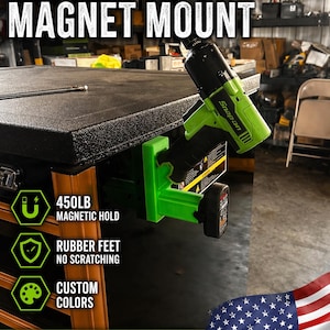 Puede incluir: Soporte magnético verde y negro de 14,4 V para herramientas y baterías. Cuenta con una sujeción magnética de 204 kg, pies de goma y opciones de color personalizadas. El texto dice "HOLDS 1 TOOL + 1 BATTERY" y "MADE IN USA".