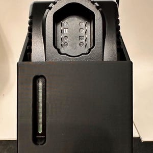 May include: A black battery charger with a rectangular base and a slotted top. The charger has a vertical indicator light on the front. The top has a gear-like design around the charging port.