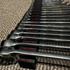 May include: A set of polished chrome wrenches, arranged in a neat row. Each wrench is labeled with SAE measurements, ranging from 1/4 to 1 1/2 inches. The tools are displayed on a textured, neutral-toned surface, highlighting their reflective surfaces.