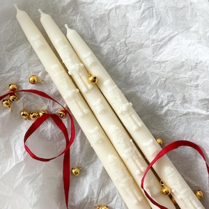 May include: Ivory-colored taper candles with embossed holiday designs, accented with gold bells and a red ribbon. The candles are arranged on a textured white surface, creating a festive and elegant display. The candles are approximately 10 inches tall.