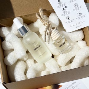 May include: An open cardboard box filled with white packing peanuts, containing two bottles of oil diffuser refill. The bottles are labeled "Amber Sage" and have silver and wooden caps. A small card with instructions is visible.
