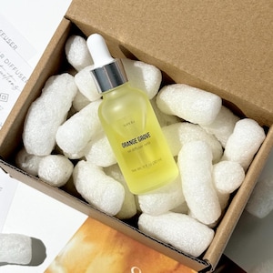 Puede incluir: Una botella de vidrio amarillo con un cuentagotas blanco y tapa plateada, etiquetada como "ORANGE GROVE oil diffuser refill", se encuentra en una caja de cartón llena de cacahuetes de embalaje blancos. La botella contiene 30 ml de líquido.