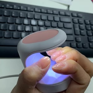 Könnte beinhalten: Eine weiße und rosafarbene Nagellampe mit einem violetten Licht, das im Inneren leuchtet. Die Lampe wird von einer Hand mit nackten Nägeln gehalten.