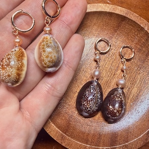 May include: Two pairs of drop earrings featuring gold-tone hoop closures. One pair showcases light brown cowrie shells with white speckles and small peach-colored pearls. The second pair features dark brown cowrie shells with white patterns and pearls.