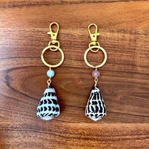 May include: Two seashell keychains with gold-colored hardware. Each keychain features a small, cone-shaped shell with a black and white pattern, a gold ring, and a small bead. The keychains are displayed on a wooden surface.