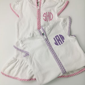 May include: Two white hooded towels with pink and purple striped trim. The towels have embroidered initials, "SHD" and "JRA".