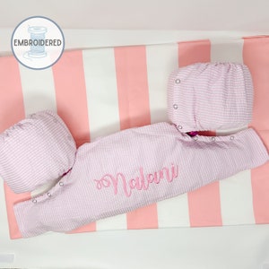 May include: A pink and white striped baby sleepsuit with the name "Nalani" embroidered on the front. The sleepsuit is laid out on a pink and white striped fabric.