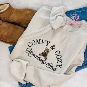 May include: Cream-colored sweatshirt with the embroidered text "COMFY & COZY Homebody Club" and a teddy bear graphic. The sweatshirt is paired with blue jeans and brown suede boots.