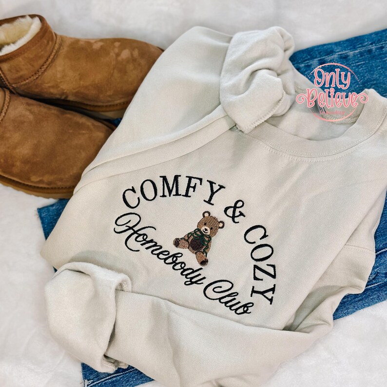 May include: Cream-colored sweatshirt with the embroidered text "COMFY & COZY Homebody Club" and a teddy bear graphic. The sweatshirt is paired with blue jeans and brown suede boots.