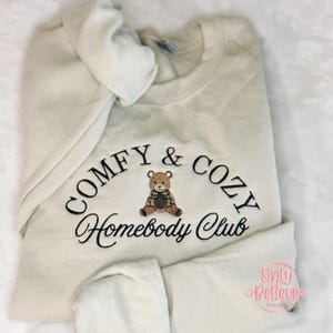 May include: Cream-colored sweatshirt with the embroidered text "COMFY & COZY Homebody Club" in black. A small teddy bear graphic is centered below the text. The sweatshirt is folded, with the hood visible.