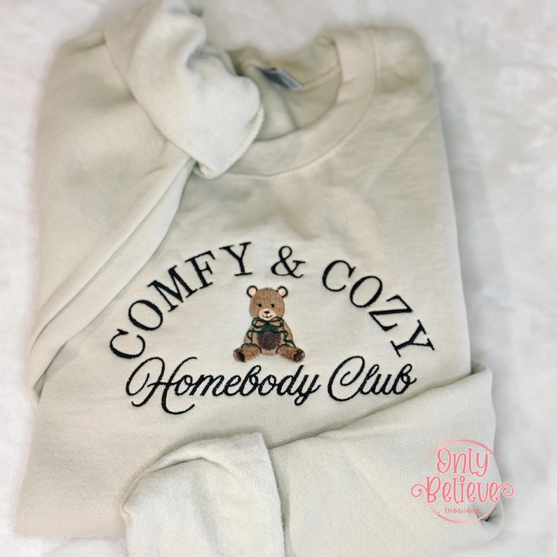 May include: Cream-colored sweatshirt with the embroidered text "COMFY & COZY Homebody Club" in black. A small teddy bear graphic is centered below the text. The sweatshirt is folded, with the hood visible.