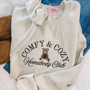 May include: A beige sweatshirt with the embroidered text "COMFY & COZY Homebody Club" and a teddy bear graphic. The sweatshirt is folded, with a portion of blue jeans and brown slippers visible.