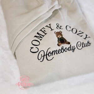 May include: A beige hoodie with the embroidered text "COMFY & COZY Homebody Club" in black. A small embroidered teddy bear wearing a green and brown plaid scarf is centered above the words "Homebody Club". The hoodie is folded on a white surface.