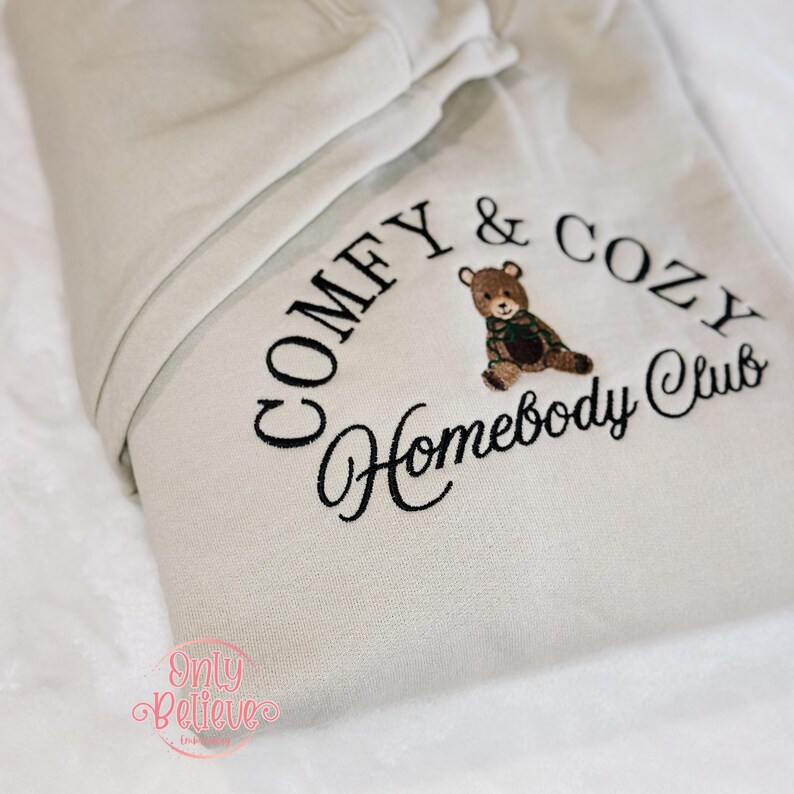 May include: A beige hoodie with the embroidered text "COMFY & COZY Homebody Club" in black. A small embroidered teddy bear wearing a green and brown plaid scarf is centered above the words "Homebody Club". The hoodie is folded on a white surface.