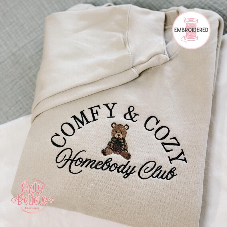 May include: A beige hoodie with the embroidered text "COMFY & COZY Homebody Club" in black script. A brown teddy bear with a green scarf is centered above the text. The hoodie has a hood and is folded.