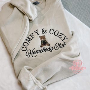 May include: A beige sweatshirt with the embroidered text "COMFY & COZY Homebody Club" in black script. A brown teddy bear with a green scarf is centered within the design. The sweatshirt has a crew neck and long sleeves.