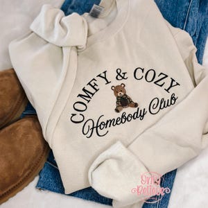 May include: Cream-colored sweatshirt with the embroidered text "COMFY & COZY Homebody Club" and a brown teddy bear graphic. The sweatshirt is folded on top of blue jeans and brown boots.